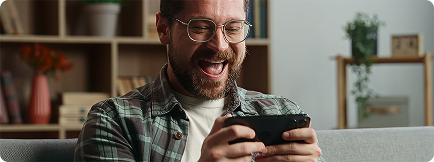 Man sitting on a couch smiling and reacting while playing a mobile game on his smartphone.
