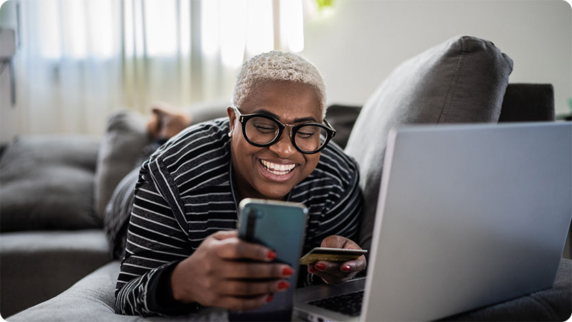 Happy person laying on a couch at home using a laptop while holding a mobile phone and a credit card.