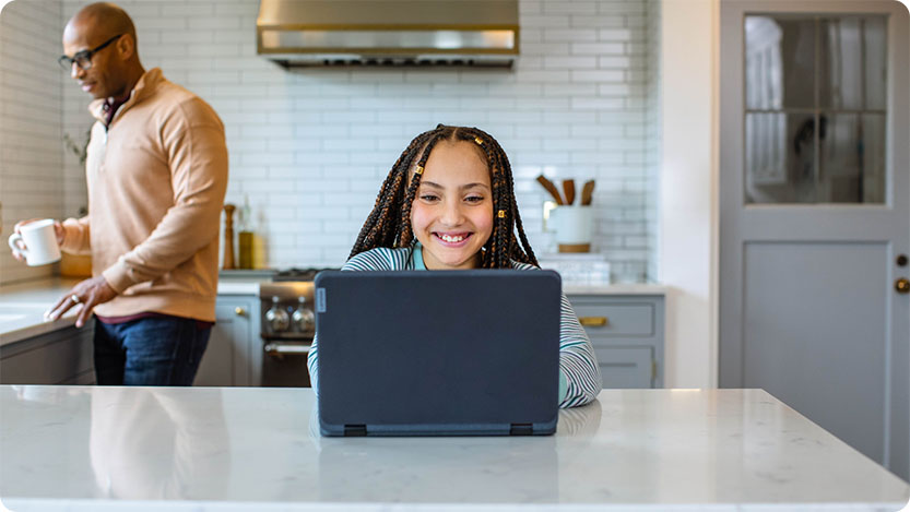 Kind mit Laptop an einer Küchentheke. Im Hintergrund geht eine Person vorbei.