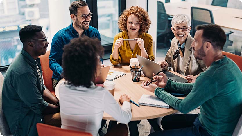 Six people sitting at a round conference table with notepads and laptops.