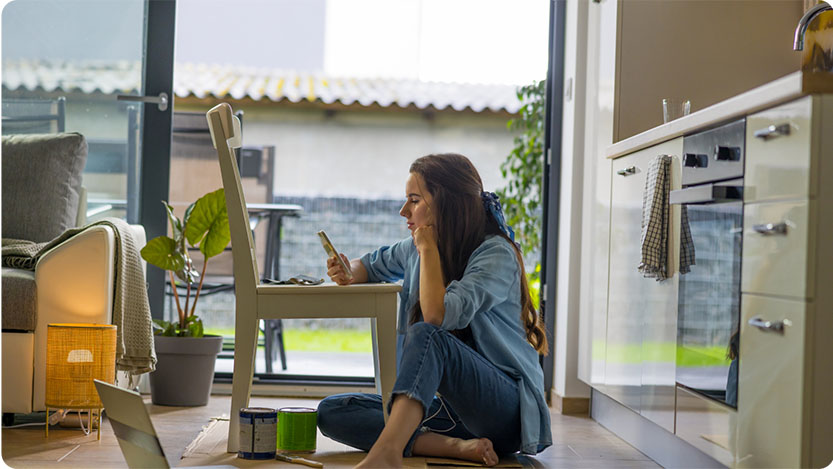 A person sitting on the kitchen floor looking at a cell phone with cans of paint and a laptop in front of them.