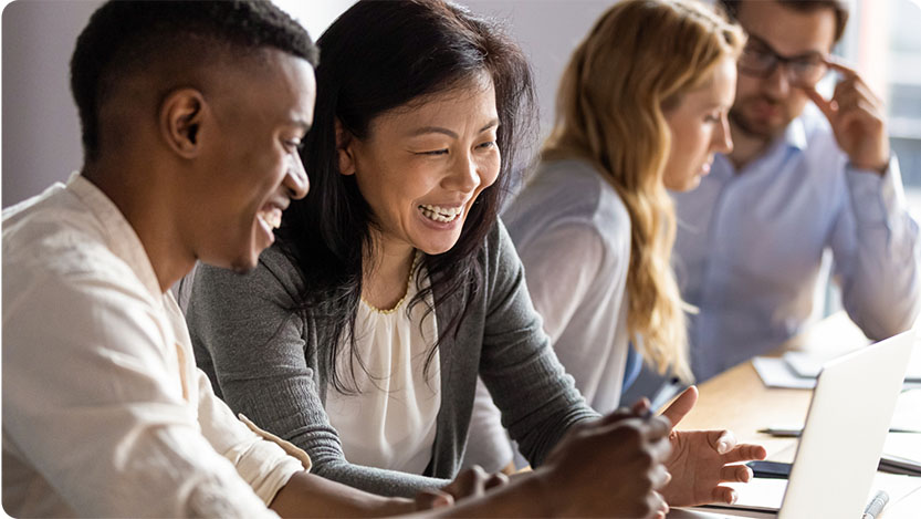 Four people sitting indoors at a desk. The two people in the front are looking at a laptop and smiling.