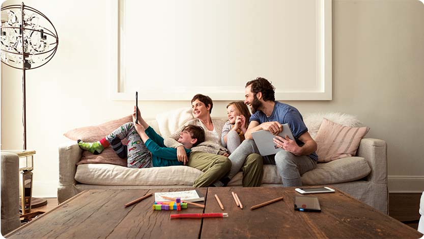 A family of four people on a couch inside a room. One person is holding a laptop. One kid is holding a tablet. Everyone is looking at the tablet and smiling.