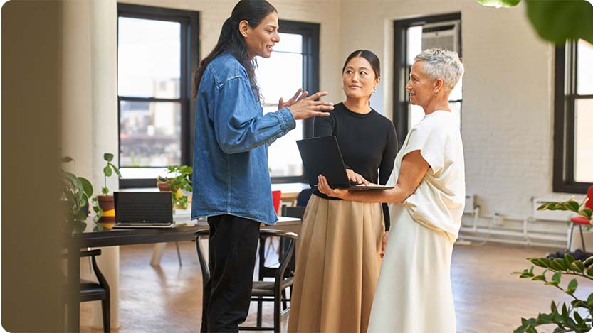 Three people standing indoors in a room and talking. One person is holding a laptop.