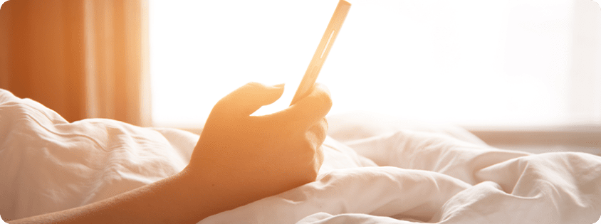 Hand holding a smartphone while lying in bed with sunlight in the background