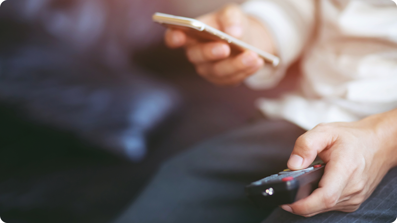 Close-up of hands holding a smartphone and a TV remote while sitting on a couch 