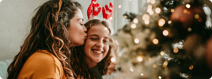 Family holiday moment by a decorated Christmas tree, with an adult and child wearing festive reindeer antlers in a warm, cozy home decorated for the holiday season.