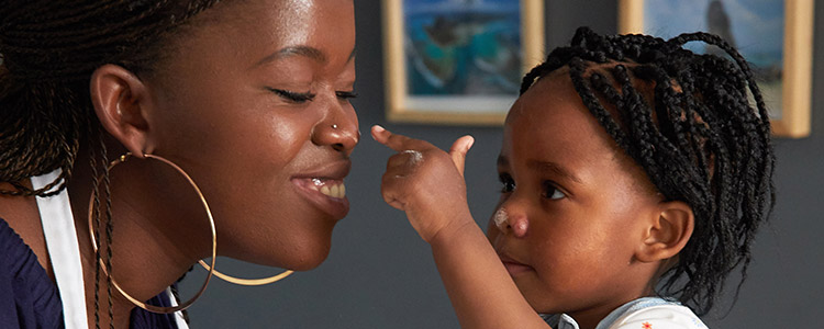 A little girl is about to touch her mother’s nose while she smiles with her eyes closed.