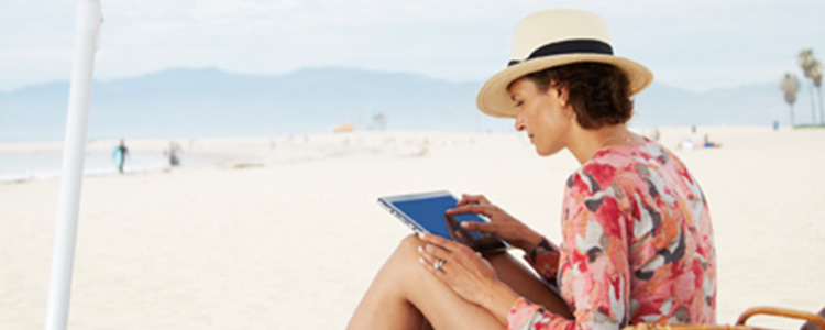 Eine Frau mit Hut sitzt am Strand mit einem Tablet.