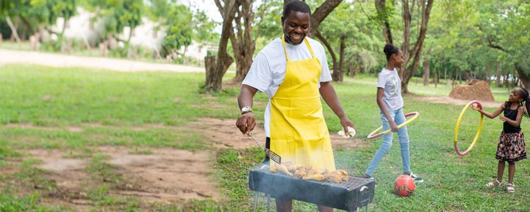 A man wearing an apron prepares a BBQ while two young girls play with hula hoops behind him.