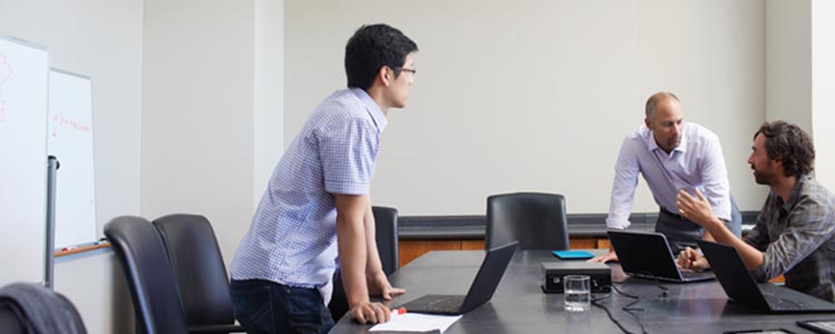 A group of three men sit in a meeting room and talk to each other while using their laptops.