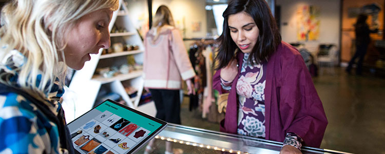 Deux femmes regardent des articles sur un comptoir tandis que l'une d'elles tient une tablette affichant un catalogue de vêtements.