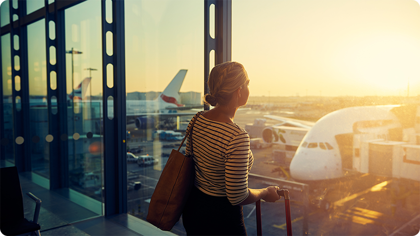 Traveler with suitcase in airport terminal at sunrise or sunset, looking out at airplanes on the tarmac through large glass windows.