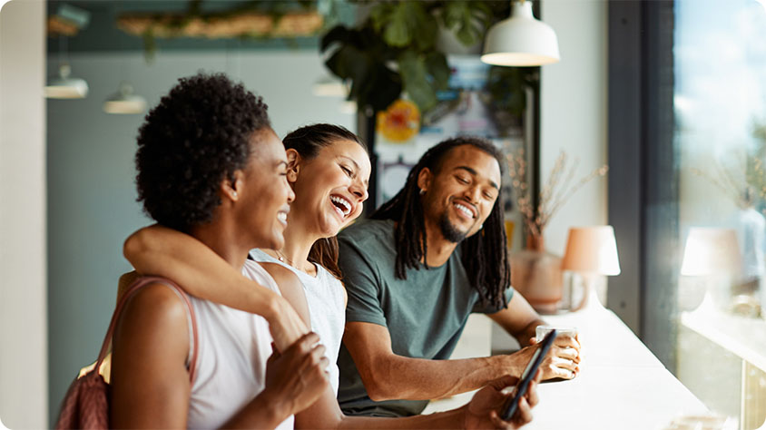 Three people sitting at a counter in a well-lit cafe. One person on the left holds a drink and has an arm around the person in the middle, who wears a white top. The person on the right, with long hair, is holding a smartphone. The background features hanging lights, plants, and large windows.