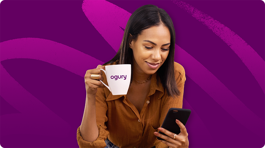 Woman checking her phone while drinking from a branded Ogury mug, representing digital engagement and brand identity.