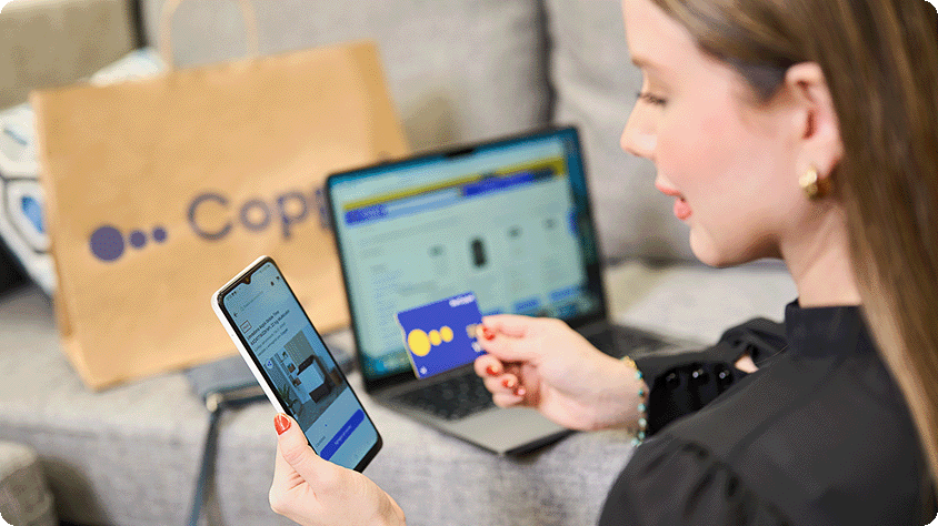 Person shopping online at home, holding a smartphone and a blue credit card in front of an open laptop, with a Coppel shopping bag in the background.