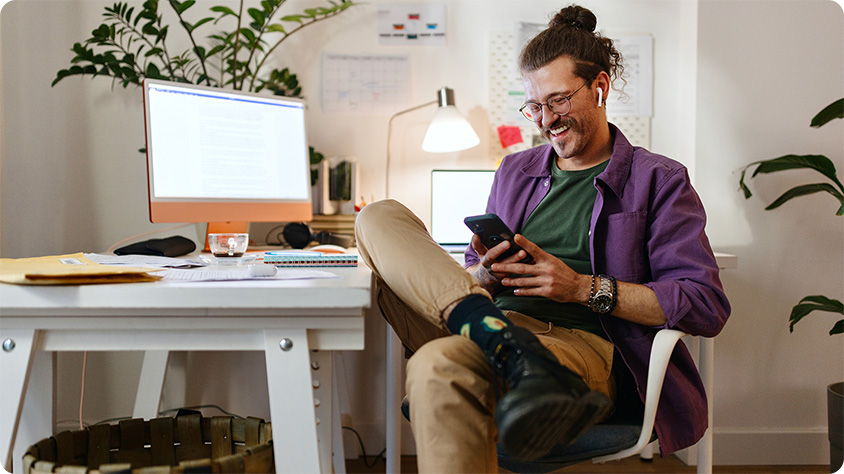 A smiling man with glasses and a bun hairstyle sits relaxed in a home office, looking at his phone while wearing wireless earbuds, with a desk, computer, and plants in the background.