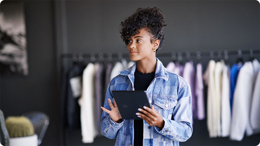 A person standing indoors holding a tablet. There is a rack with jackets in the background.