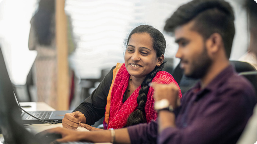 Two people sitting indoors at a desk, working on computers.