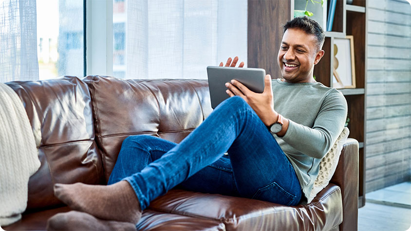 A person is sitting on a brown leather couch in a modern living room, holding and using a tablet.