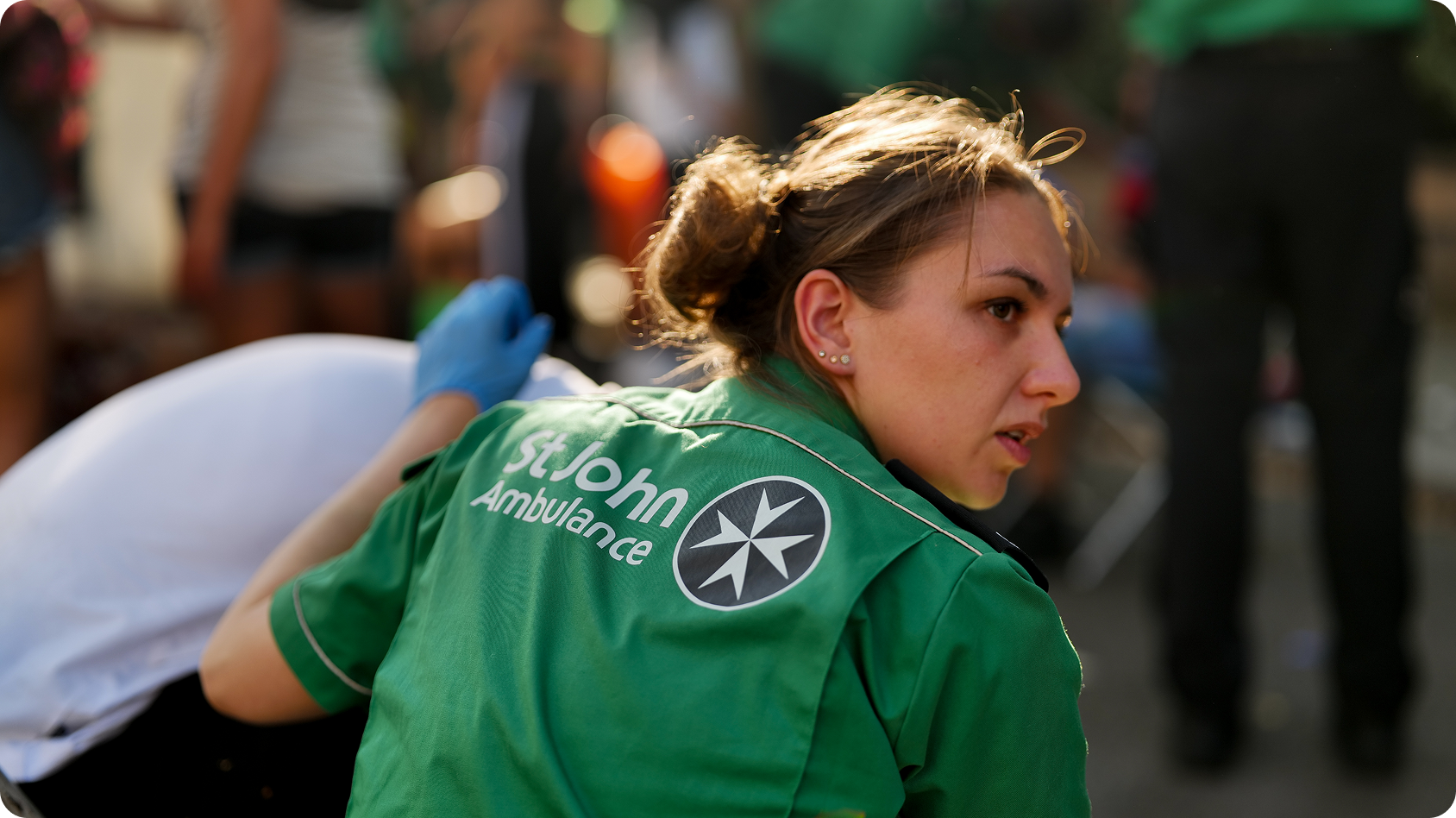  A person in a green 'St John Ambulance' shirt attends to someone lying down, with blurred background figures and objects.