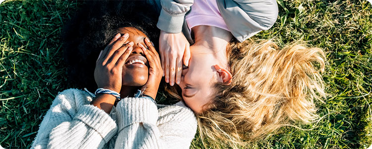 Zwei Menschen, die im Gras liegen. Der eine Mensch flüstert dem anderen Menschen etwas ins Ohr.