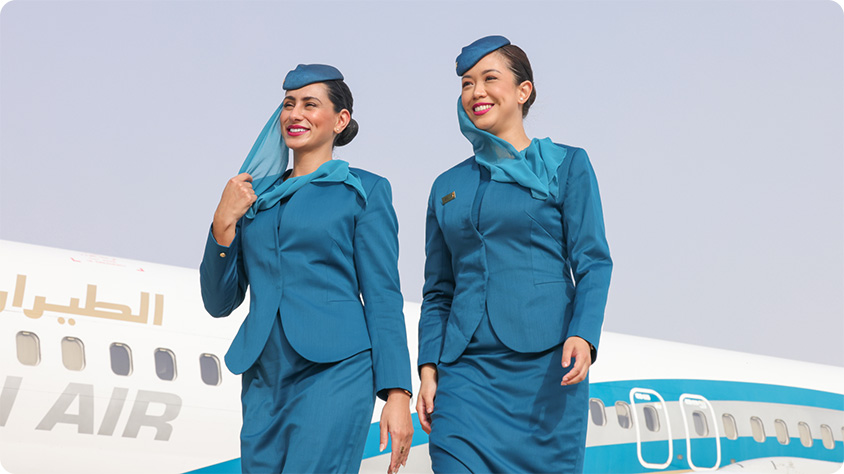 Two flight attendants wearing teal Oman Air uniforms, including jackets, skirts, and matching hats, are walking in front of an airplane. The airplane has Arabic text and the words 'AIR' visible on its side. The background shows a clear sky.